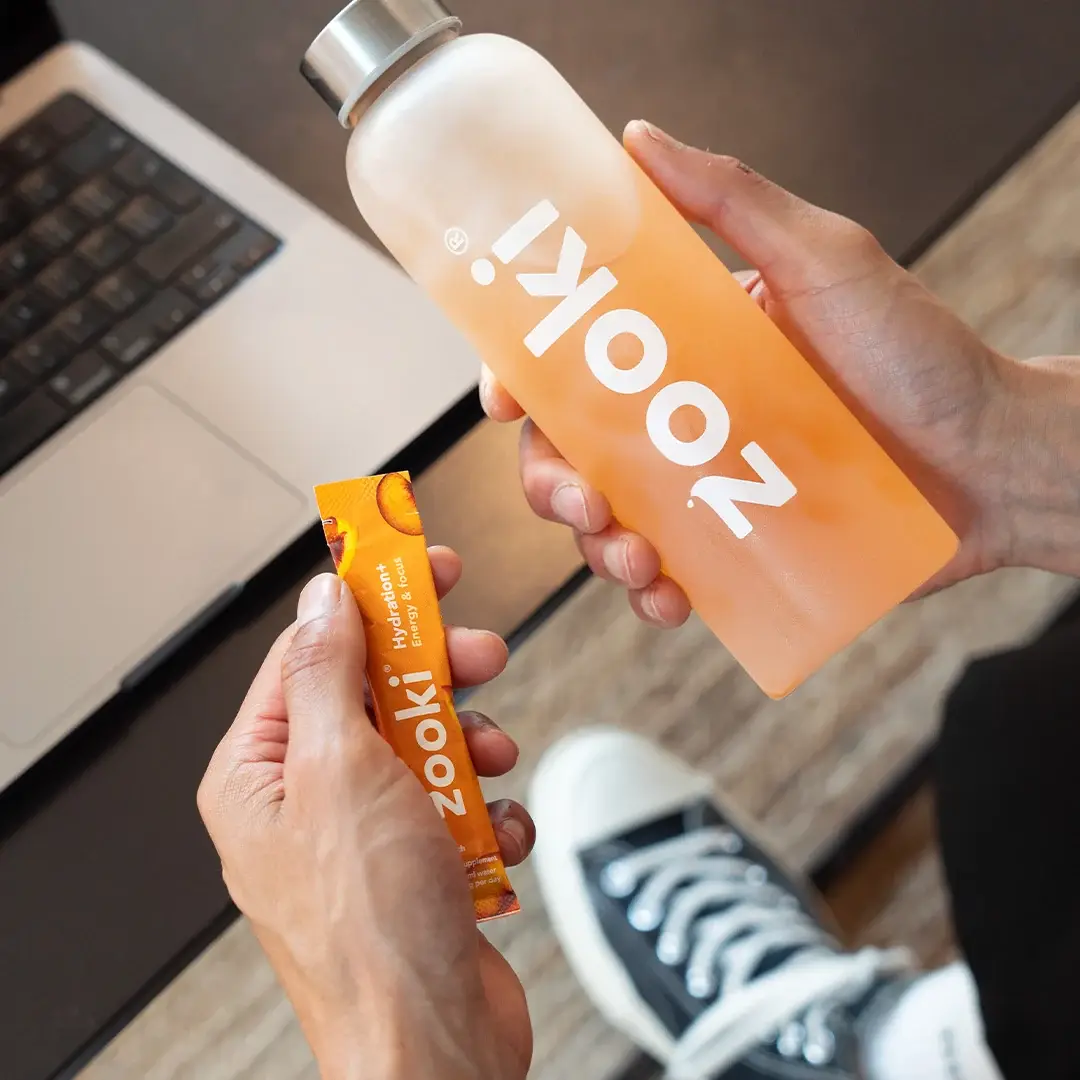 Hands holding a Zooki bottle and sachet over a laptop keyboard